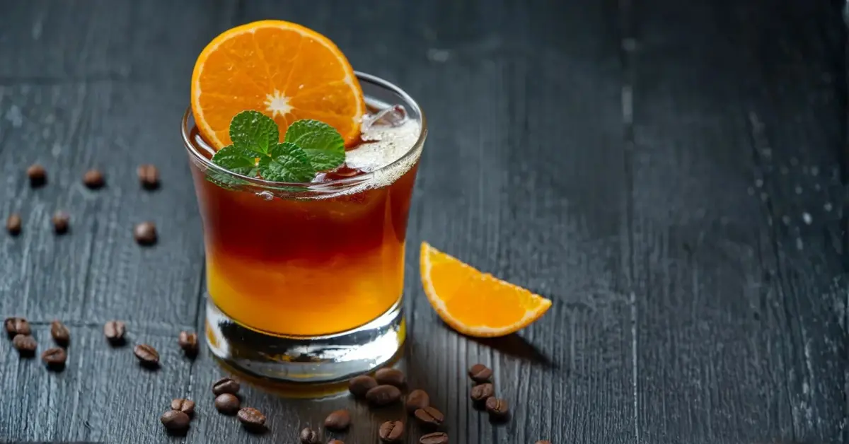 Warstwowy koktajl kawowo-pomarańczowy w szklance z lodem, dekorowany plastrem pomarańczy i miętą.